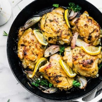 bone-in chicken thighs with crispy skin in skillet with lemon slices and onions