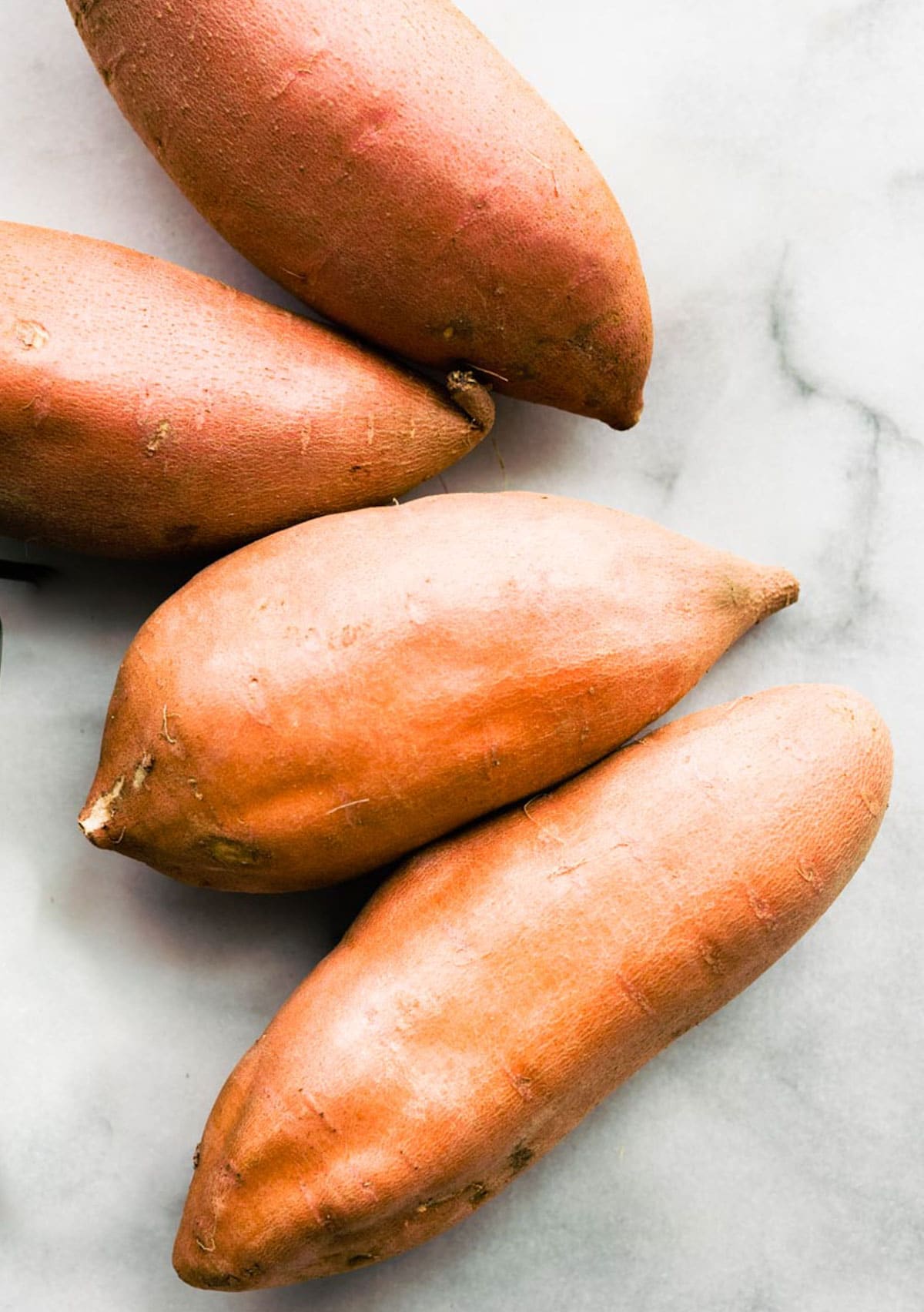 Four sweet potatoes with skin on laying on marble countertop together.