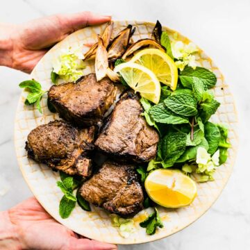 a hand holding a plate with lamb chops, fresh mint, and sliced lemon