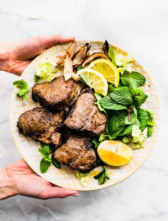 a hand holding a plate with lamb chops, fresh mint, and sliced lemon