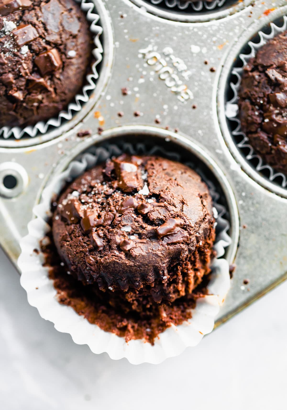 A dark chocolate vegan muffin with white liner pulled back from edges, placed halfway into muffin tin well.