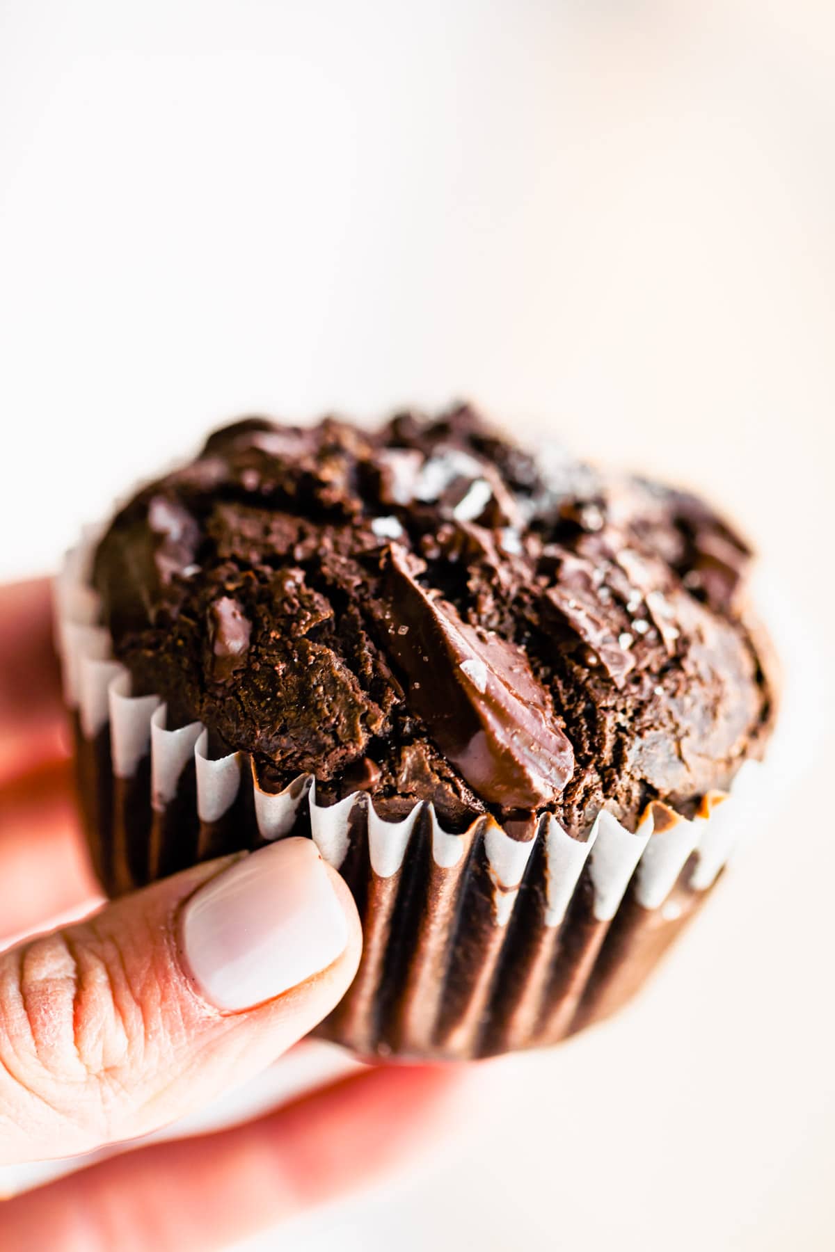 A hand holding a dark chocolate vegan muffin in white liner topped with dark chocolate chunks and sea salt flakes.