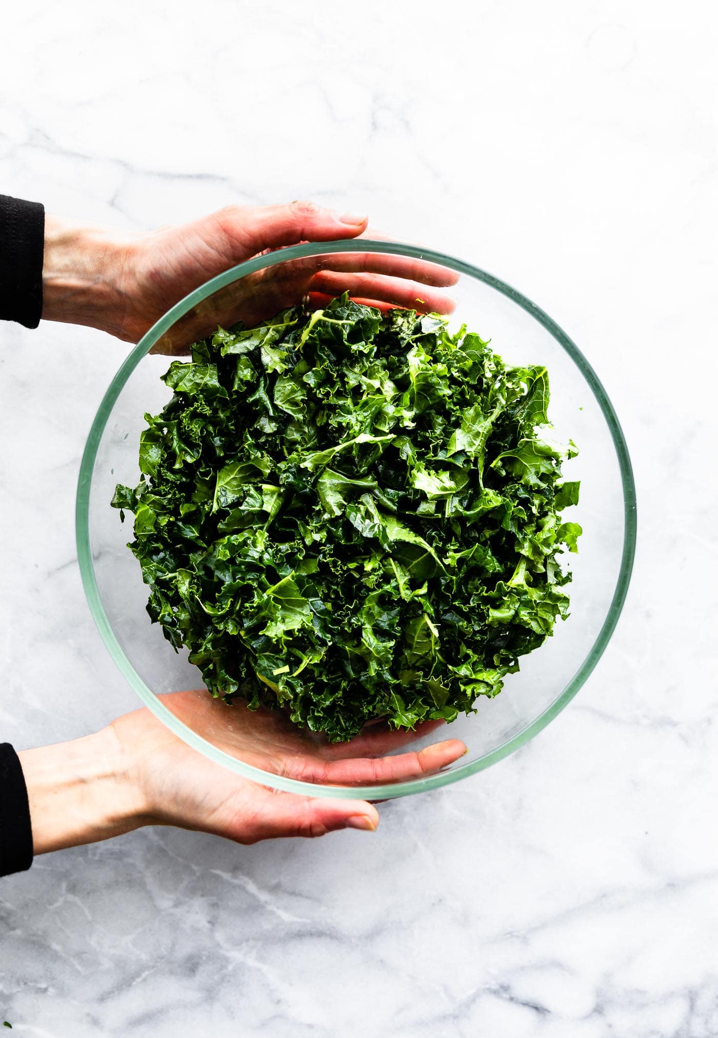 Two hands holding a clear glass bowl filled with massaged kale.