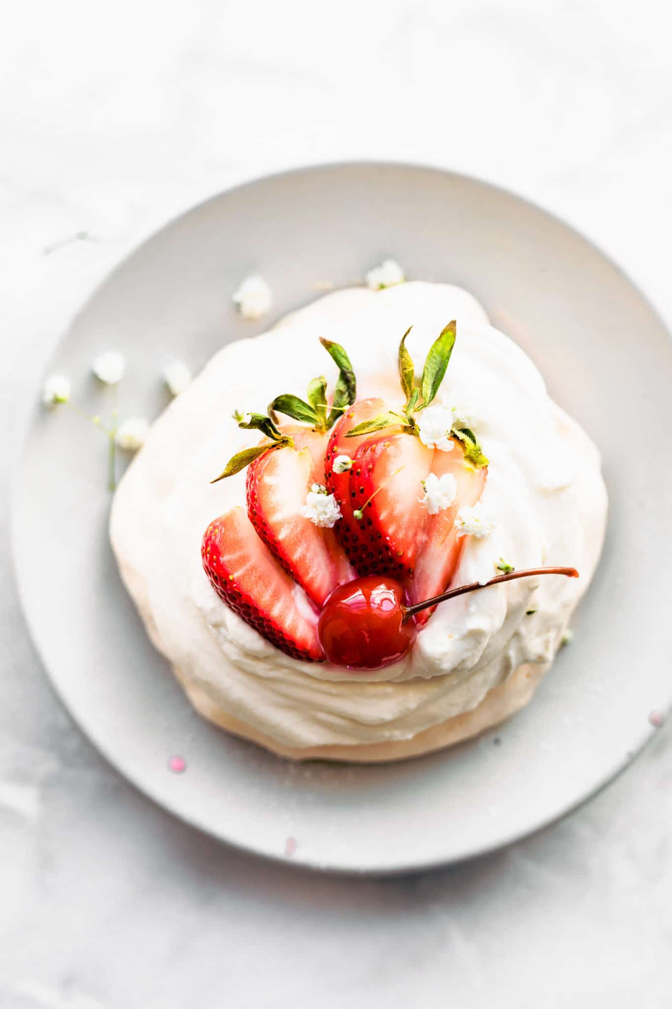An overhead image of a pavlova on a white plate, topped with fresh strawberries and a cherry.