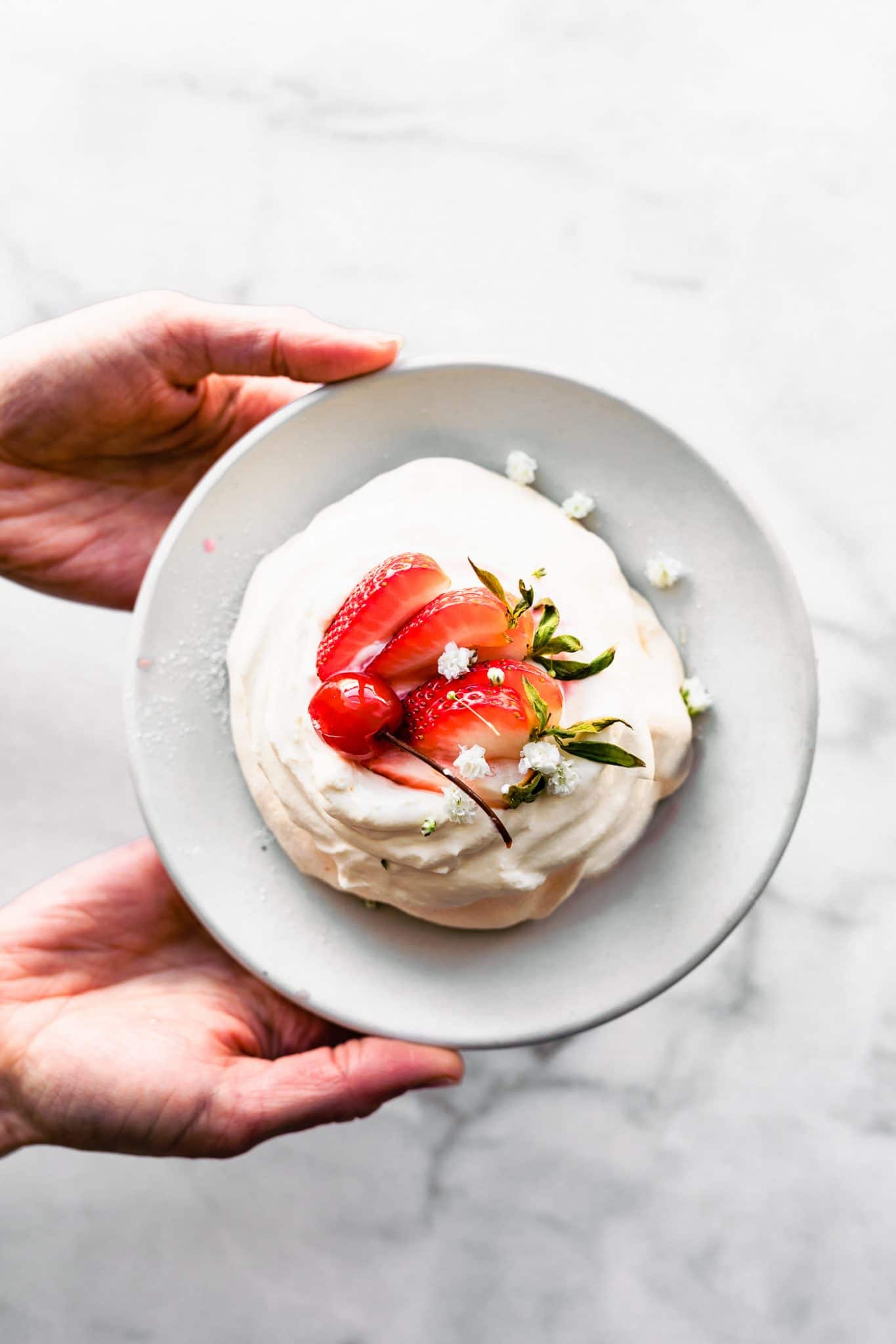 Overhead image of two hands holding a plate with a strawberry topped pavlova.