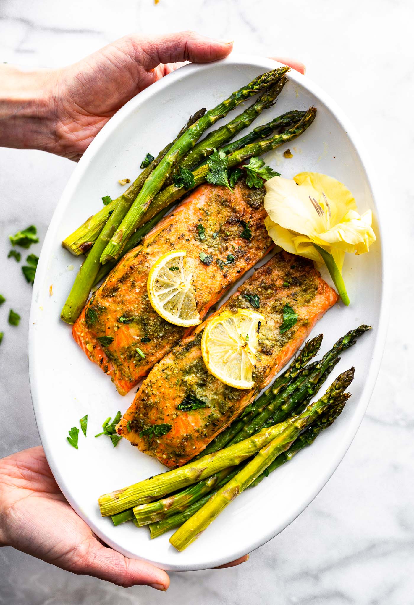 Two hands holding white platter with baked salmon filets and roasted asparagus spears, flower on the side.