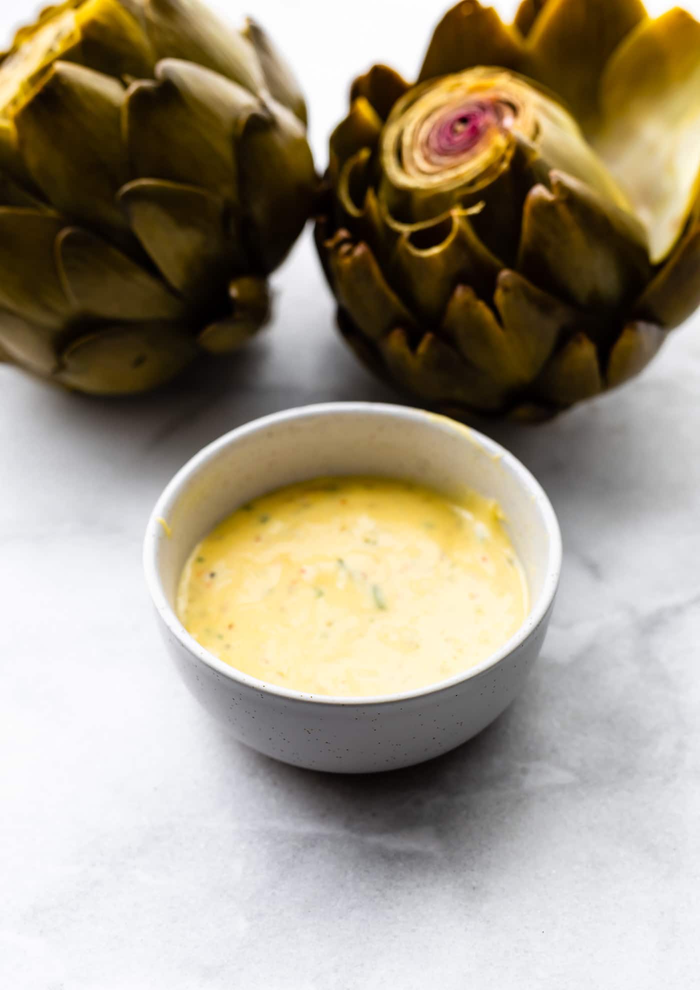 Two cooked artichokes next to a bowl of garlic aioli.