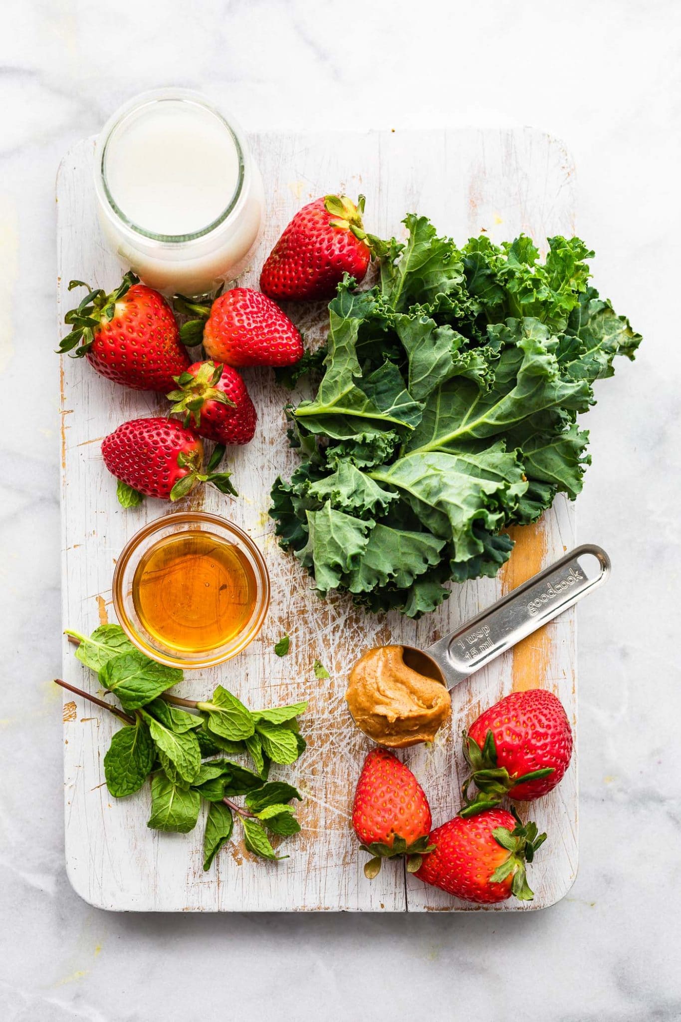 overhead photo of strawberries, kale, honey, milk, almond butter on a spoon, and mint.