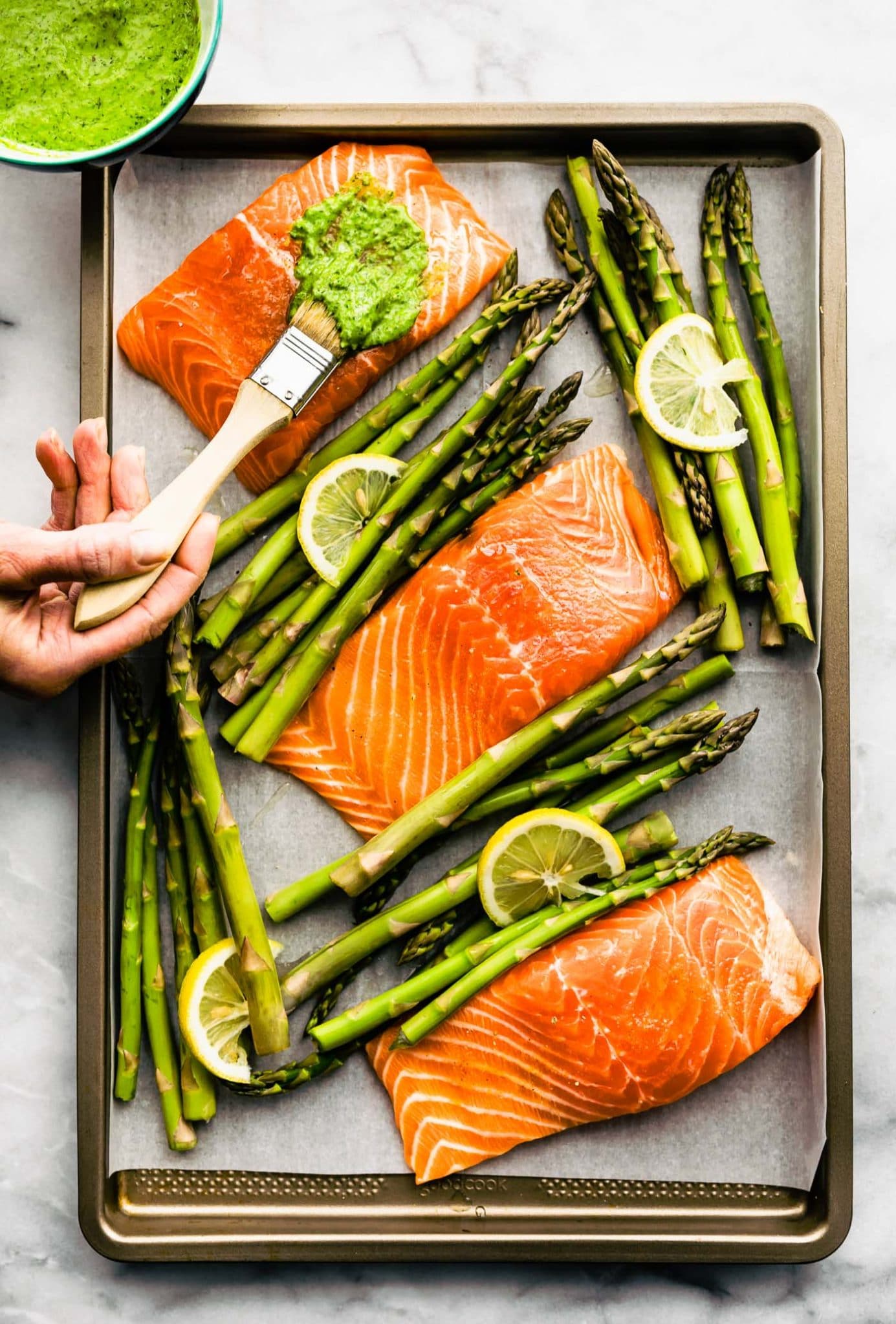 A pastry brush brushing herb sauce on raw salmon filet; baking sheet with three salmon filets and asparagus spears.