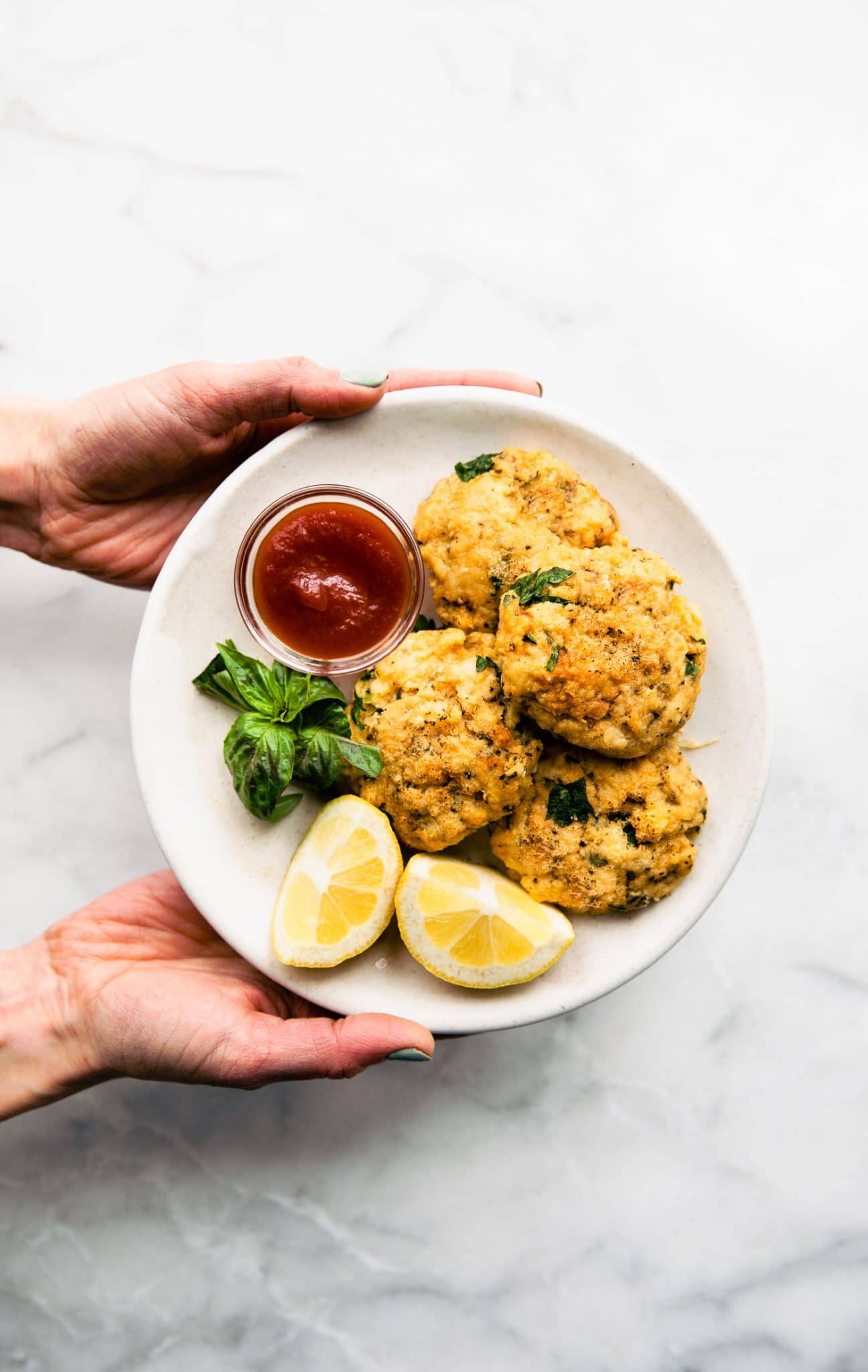 An overhead image of hands holding a plate of cod cake fish patties with fresh basil, lemon wedges, and marinara sauce.