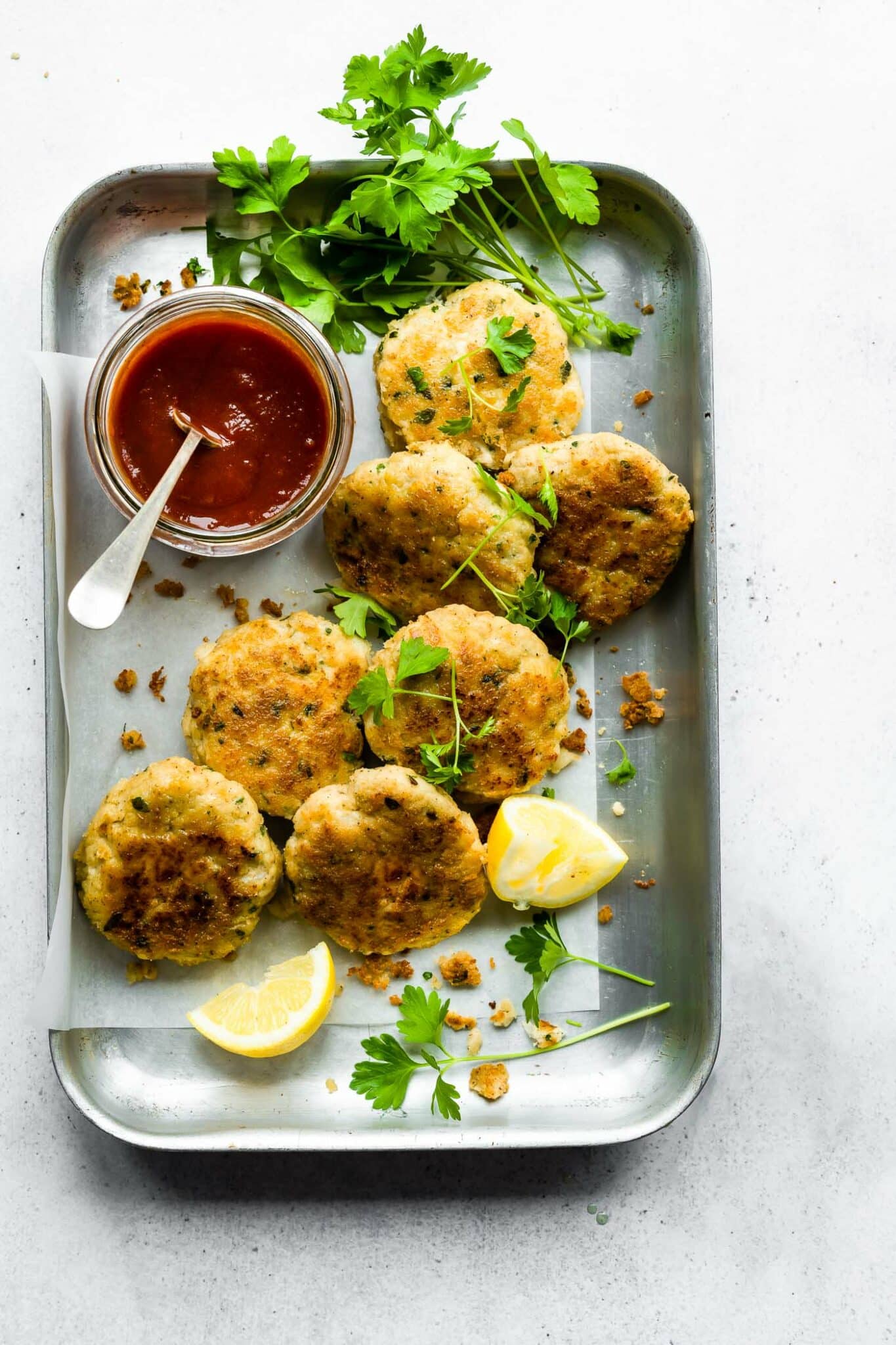 Overhead image of cod cakes layered on a tin casserole dish with a jar of marinara for dipping, and garnished with parsley and fresh lemons.