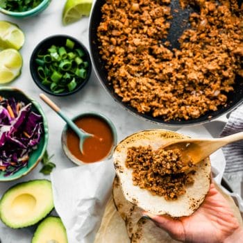 Overhead tabletop image of vegan tacos being prepared with vegan taco meat. Taco toppings including avocado, slaw, limes, cilantro, and green onions can also be seen.
