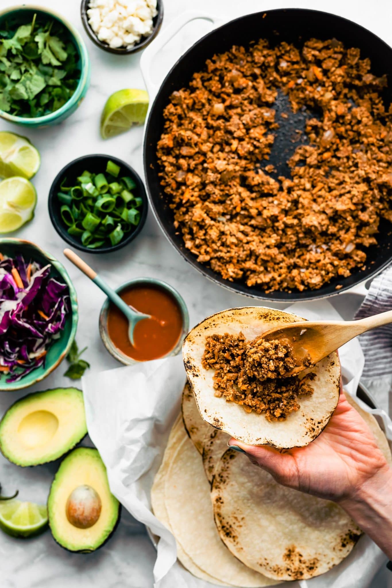 Overhead tabletop image of vegan tacos being prepared with vegan taco meat. Taco toppings including avocado, slaw, limes, cilantro, and green onions can also be seen.