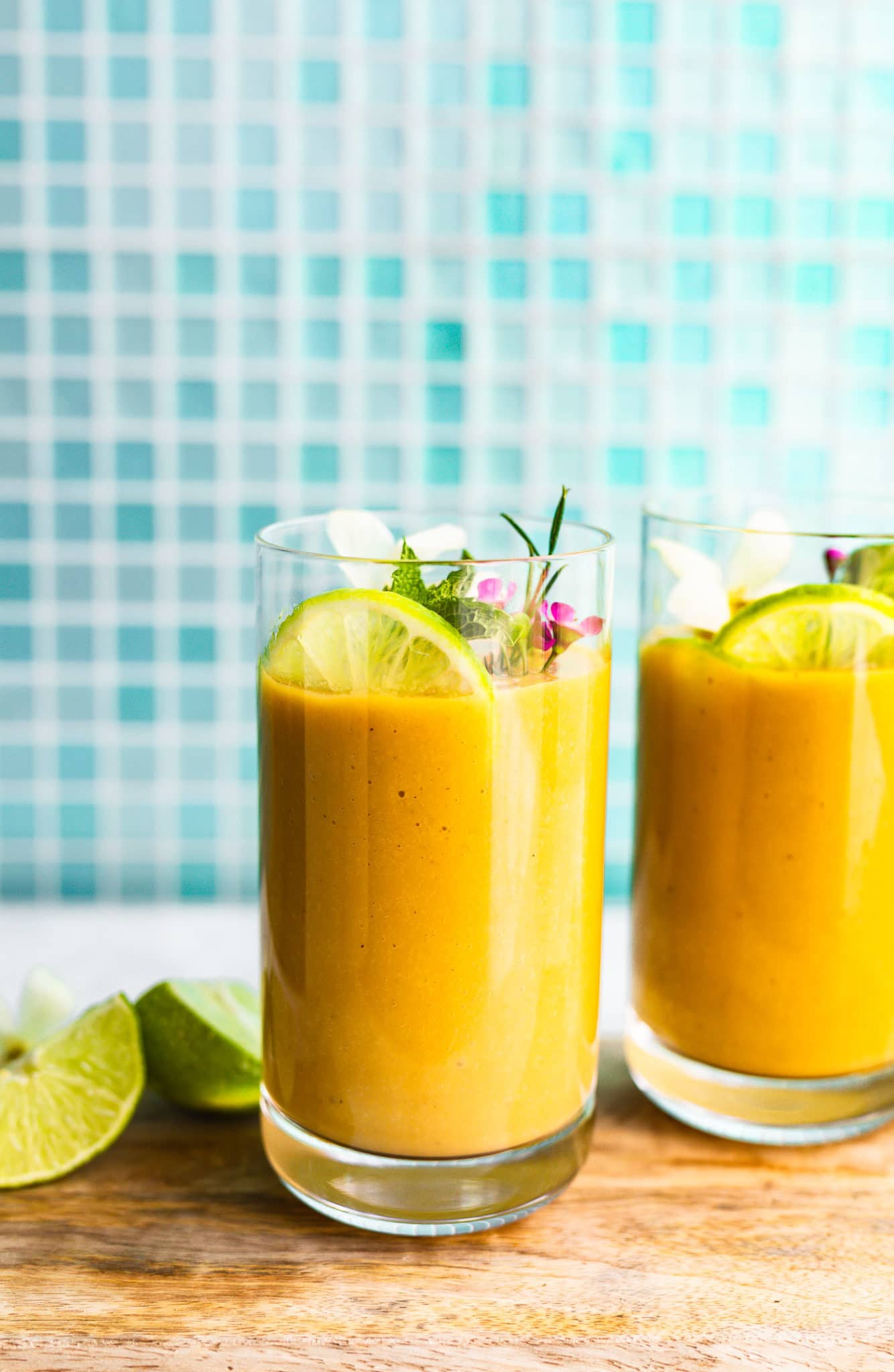 A side shot of two turmeric smoothies in glasses on a serving tray with a garnish of lime, mint, and flowers.