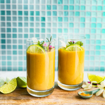 A side shot of two turmeric smoothies in glasses on a serving tray with a garnish of lime, mint, and flowers.
