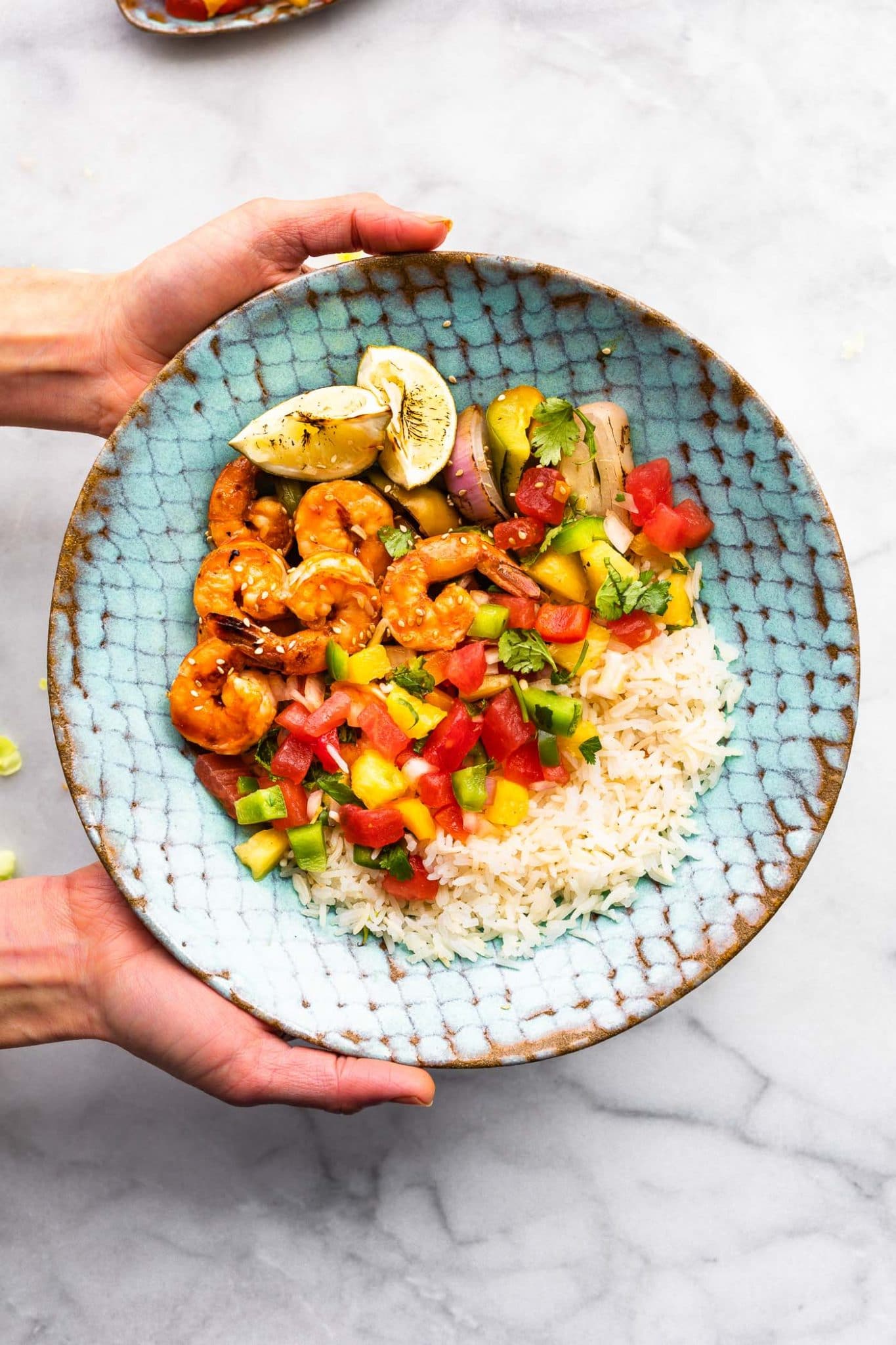 Two hands holding blue pottery plate with serving of white rice, chile-ginger marinated grilled shrimp and watermelon salsa.