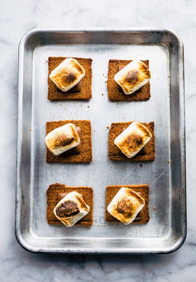Six gluten free graham crackers with toasted homemade marshmallow lined up on a baking sheet.