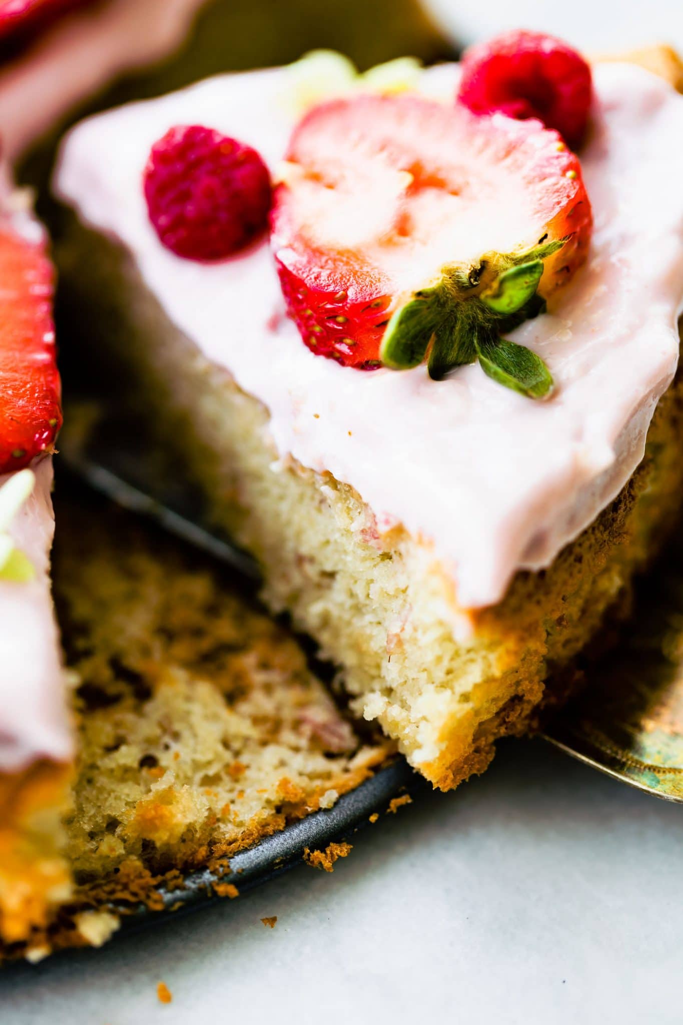 Close up image of a slice of almond cake with yogurt frosting being pulled from the pan, garnished with berries.