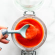Overhead image of a spoonful of homemade sriracha sauce being lifted out of a storage jar.