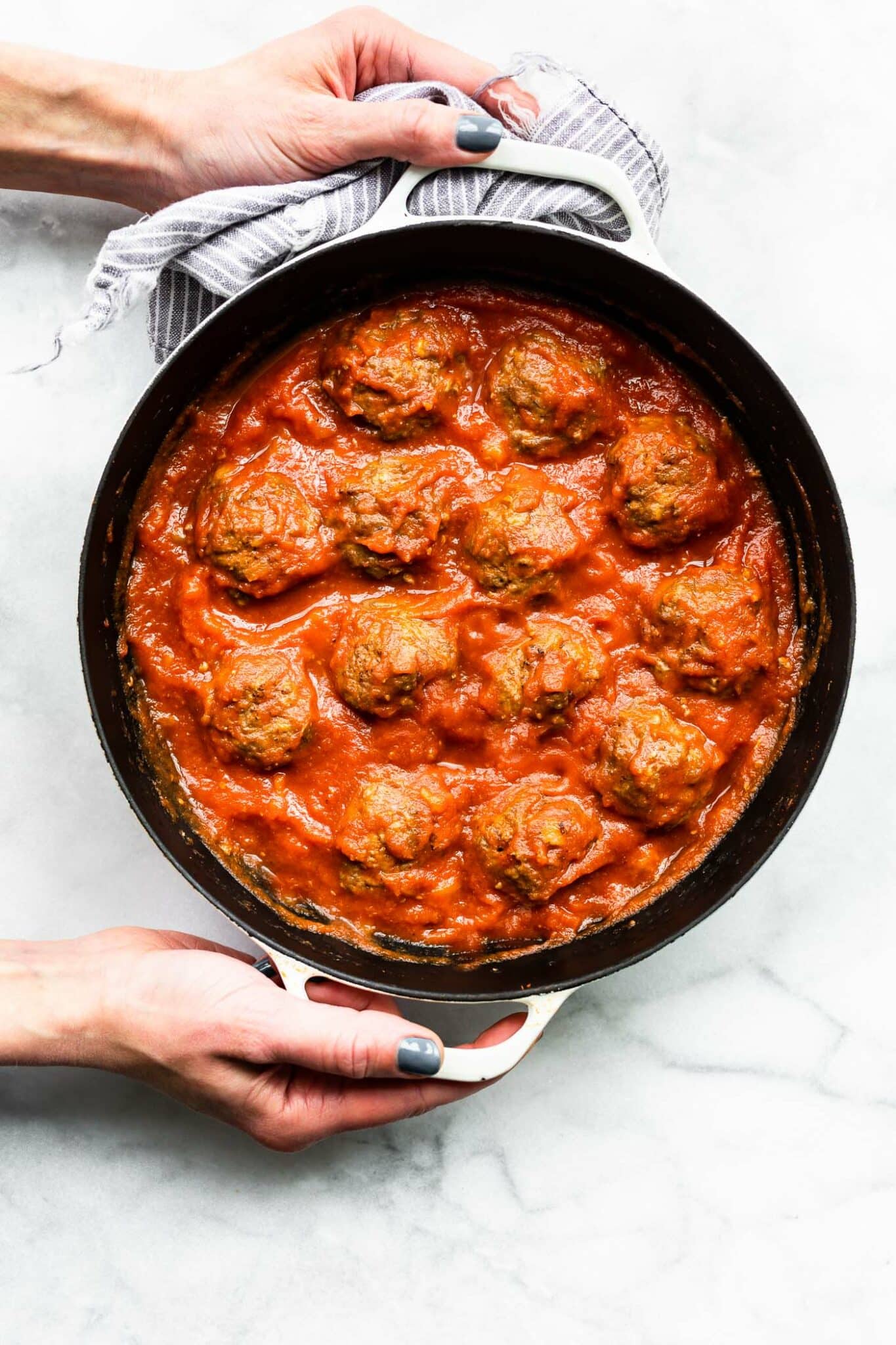 Overhead image of honey sriracha meatballs in a skillet, held by two hands over marble countertop.
