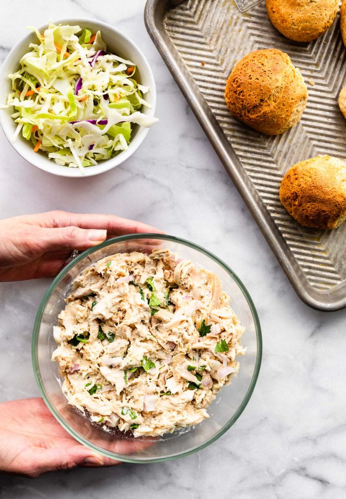 Two hands holding glass bowl with shredded chicken salad for sliders.