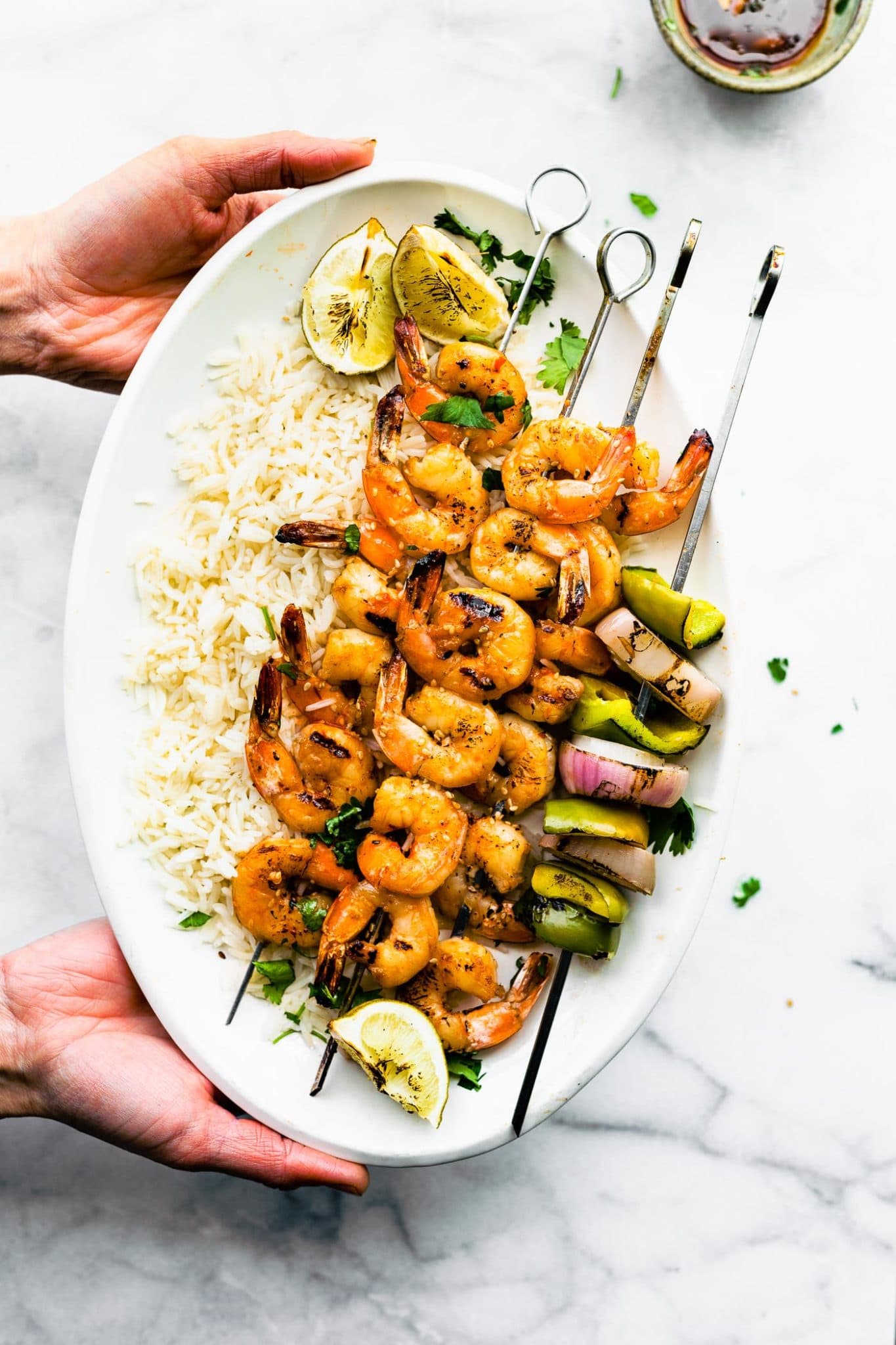 Two hands holding white platter filled with chile ginger marinated shrimp on skewers, grilled, white rice on platter.