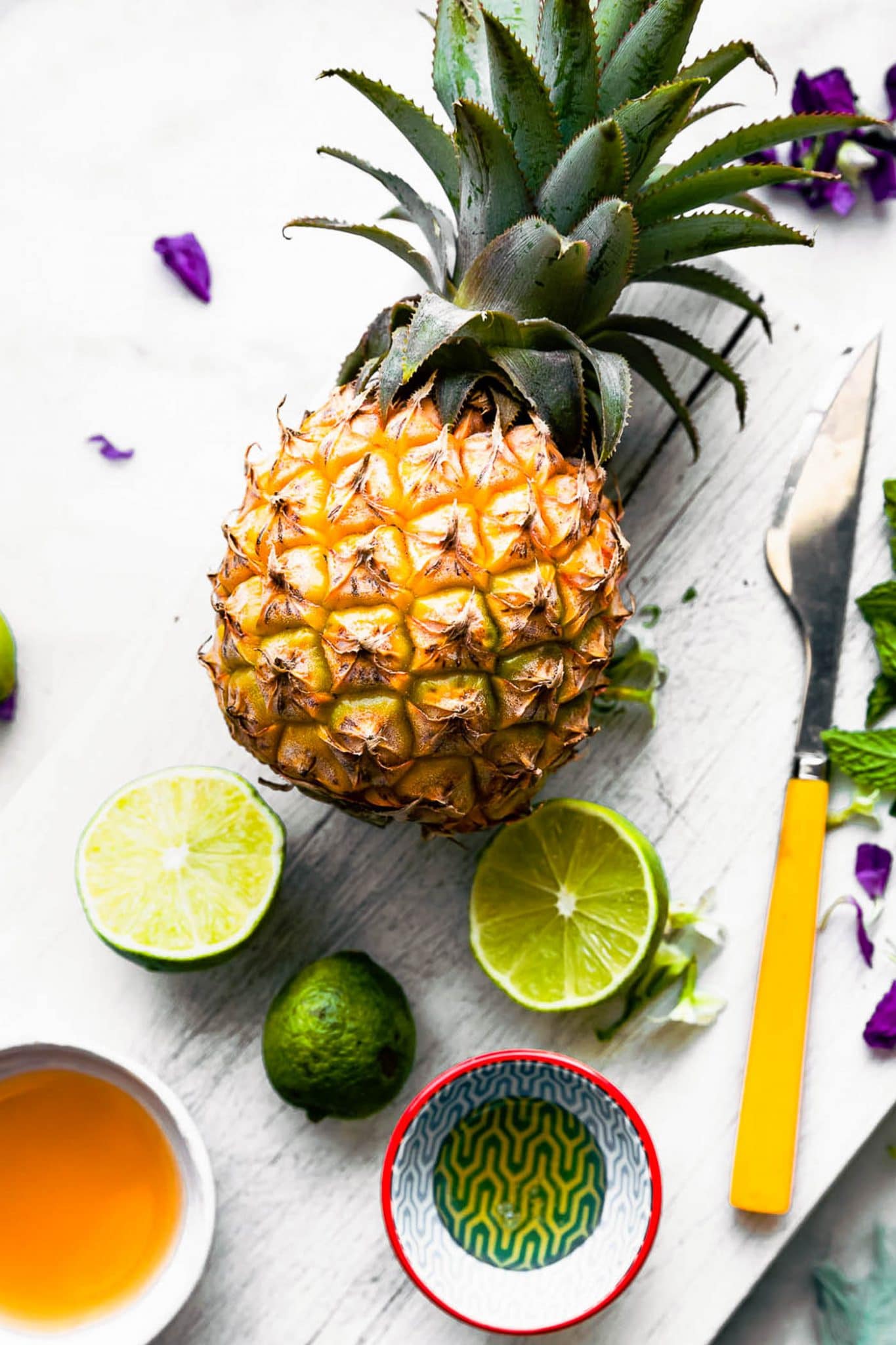 A fresh pineapple, cut limes, two bowls with liquid ingredients for pineapple watermelon salsa on white cutting board.