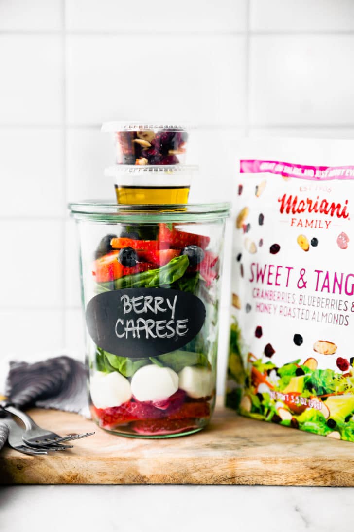 A mason jar layered with Berry Caprese salad with small container with oil and small container with salad toppings stacked on top of mason jar.