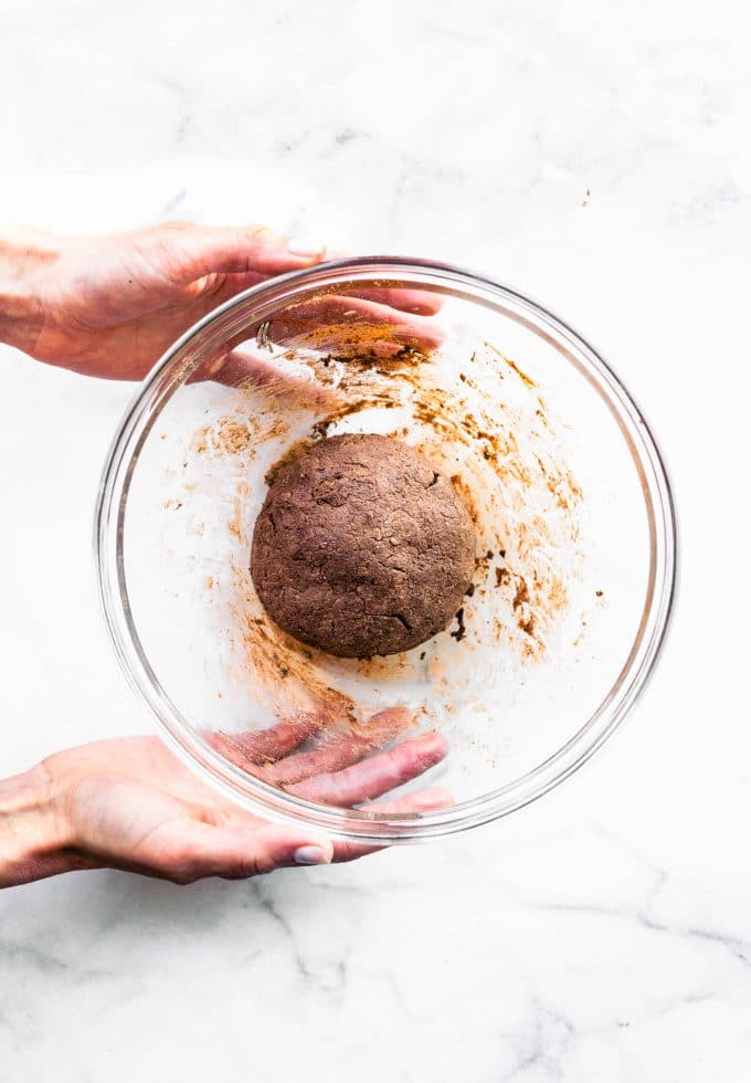 Two hands holding clear glass mixing bowl with chocolate protein cookie dough ball in center of bowl.