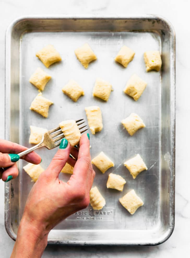 A woman's hands holding a small cauliflower gnocchi piece pressing a fork into top to make marks, over a baking sheet filled with gluten free cauliflower gnocchi pieces.