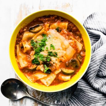 lasagna soup in a yellow bowl sitting on a board next to a spoon and blue and white towel