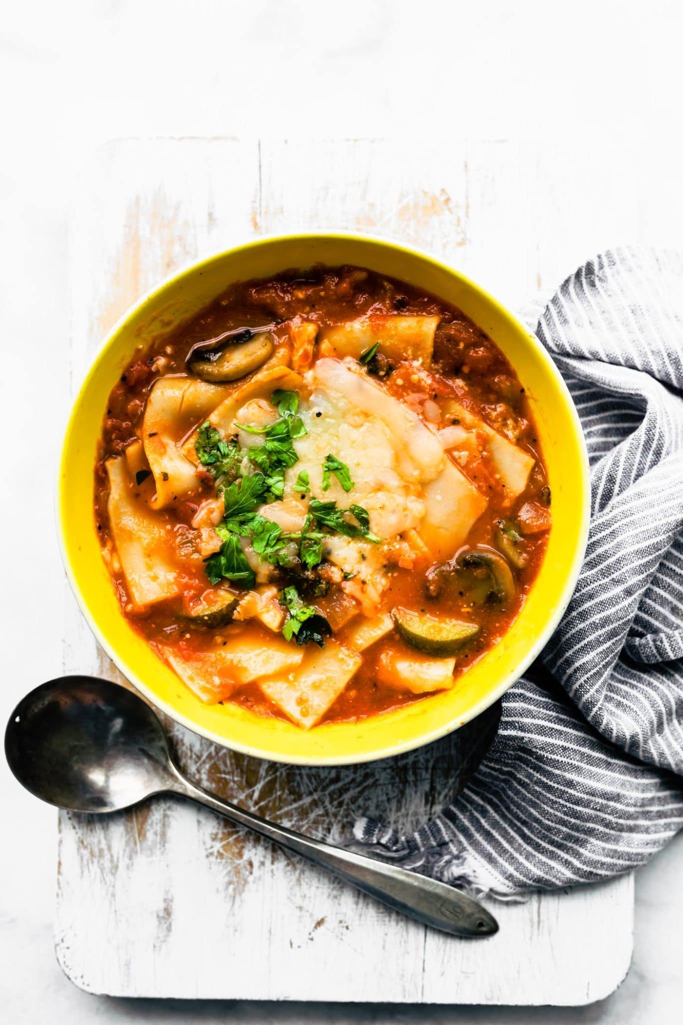 lasagna soup in a yellow bowl sitting on a board next to a spoon and blue and white towel