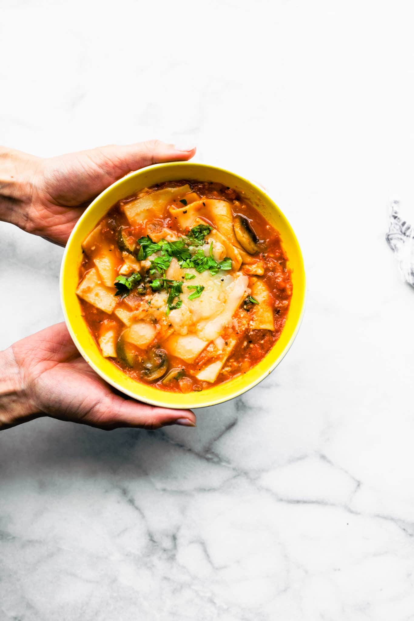 lasagna soup topped with cheese and fresh herbs being held by two hands