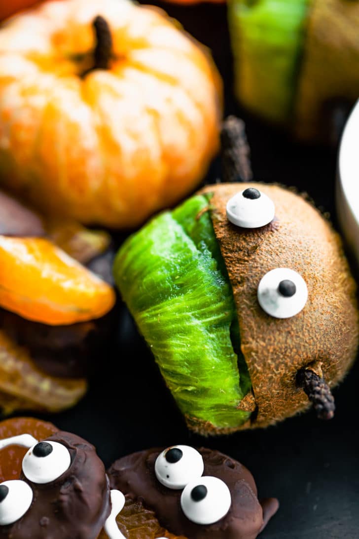 Close up view of a fresh kiwi cut to look like a frankenstein with edible google eyes for a halloween charcuterie board.