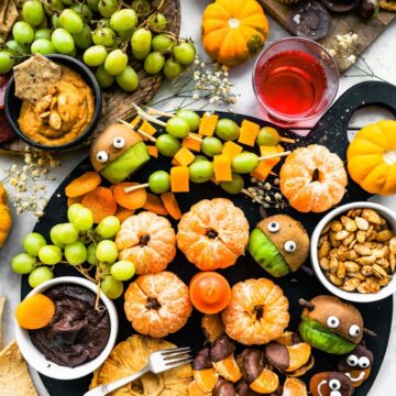 overhead photo of halloween snack board on black cheese board. Fruit, chocolates, grapes, pumpkin, and cupcakes. Dips in bowls