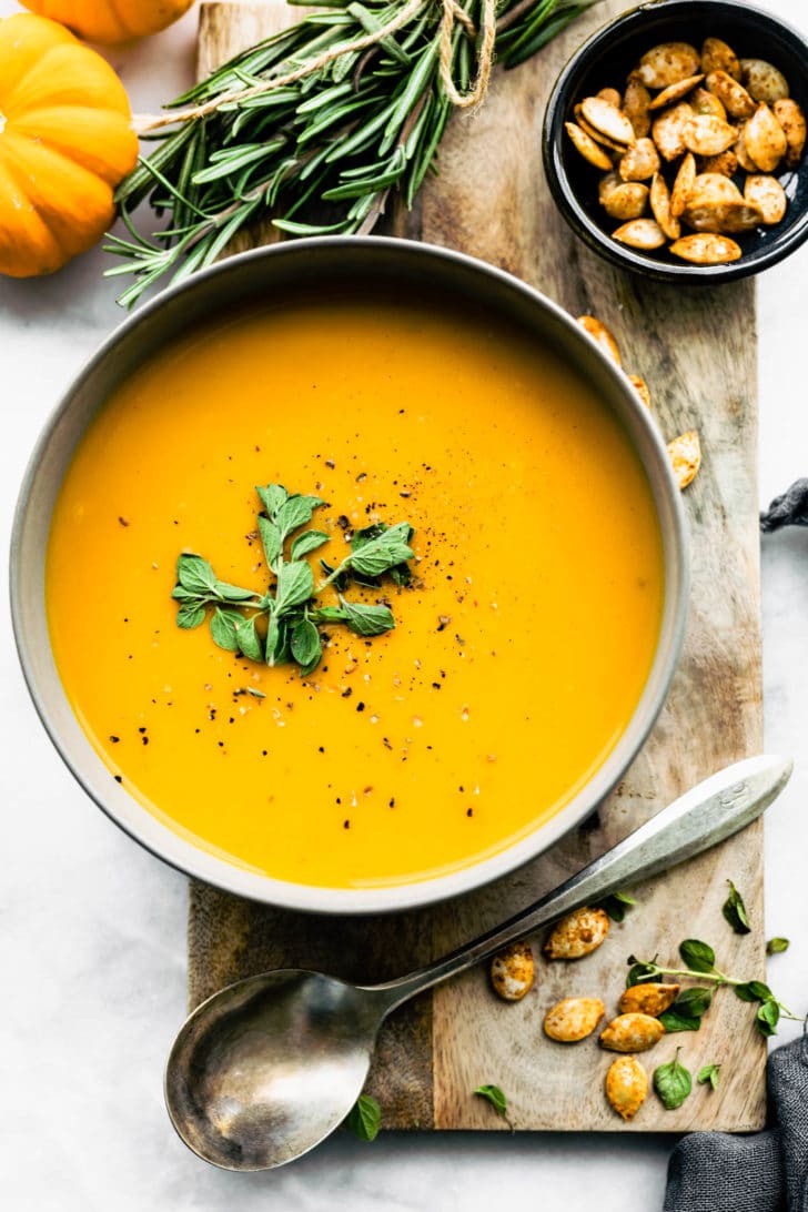 butternut squash soup served in a white bowl, surrounded by mini pumpkins, thyme, roasted pumpkin seeds, and a spoon