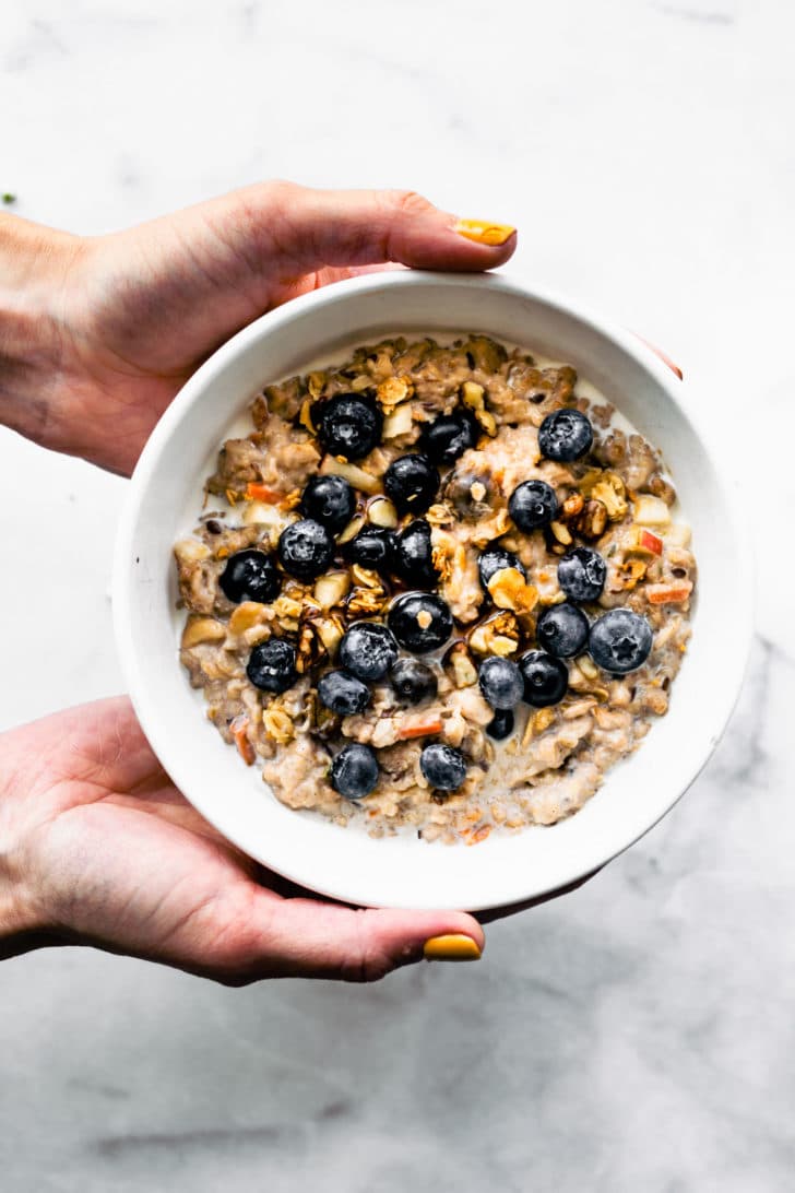 two hands holding a white bowl full of superfood oatmeal topped with fresh blueberries