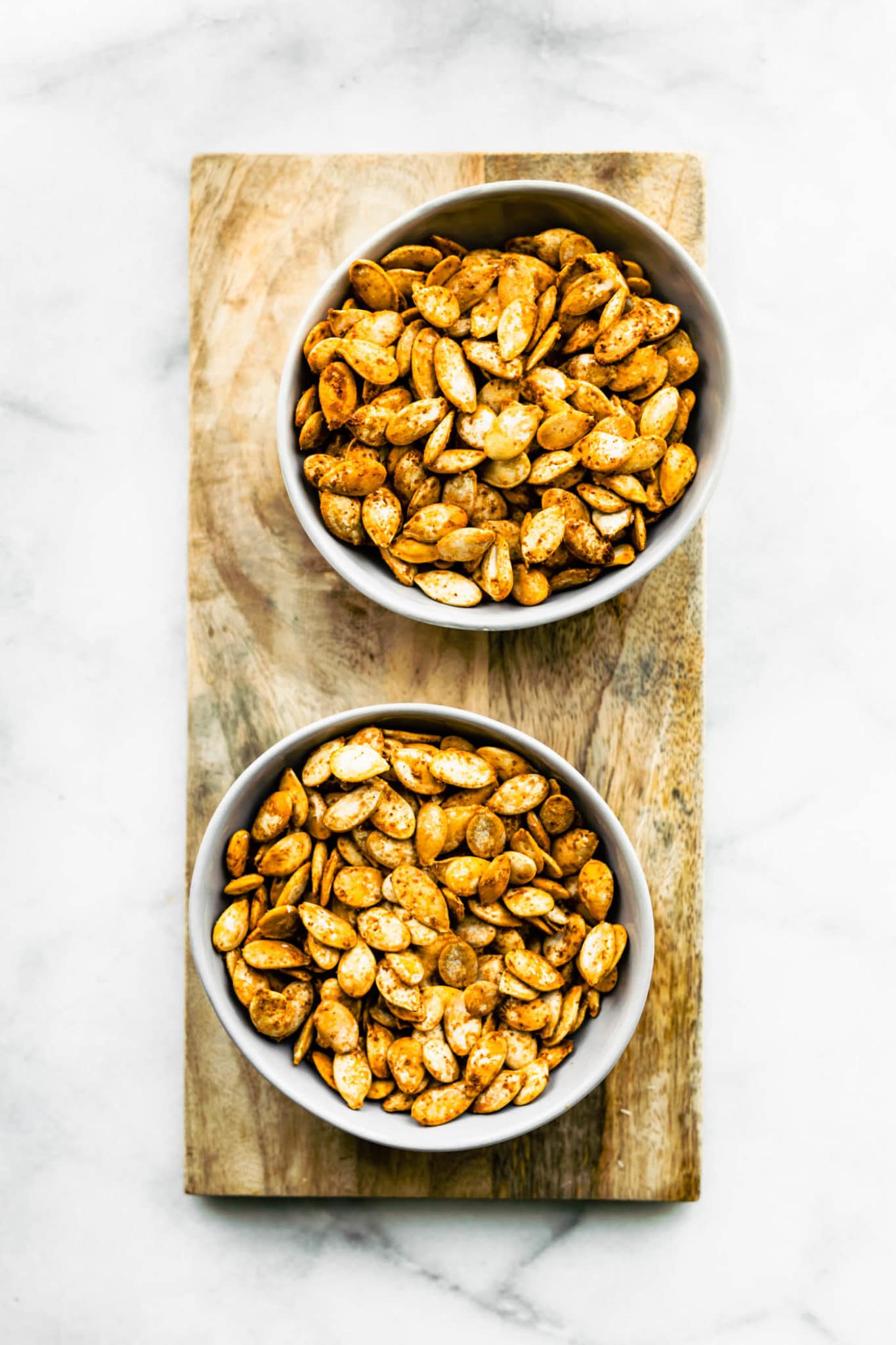 Two bowls of flavored roasted pumpkin seeds on a wooden cutting board.