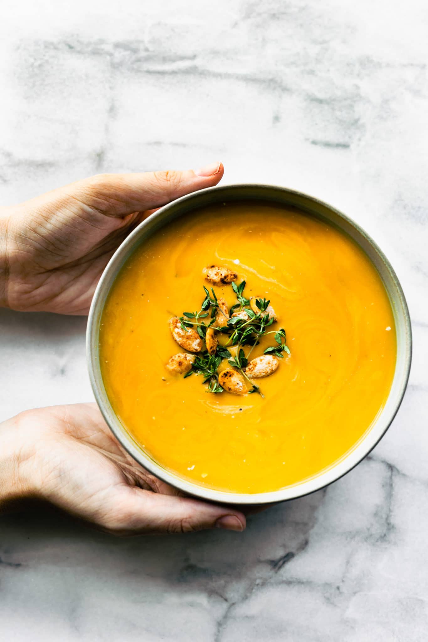 Two hands holding a bowl of butternut squash soup topped with thyme and roasted pumpkin seeds