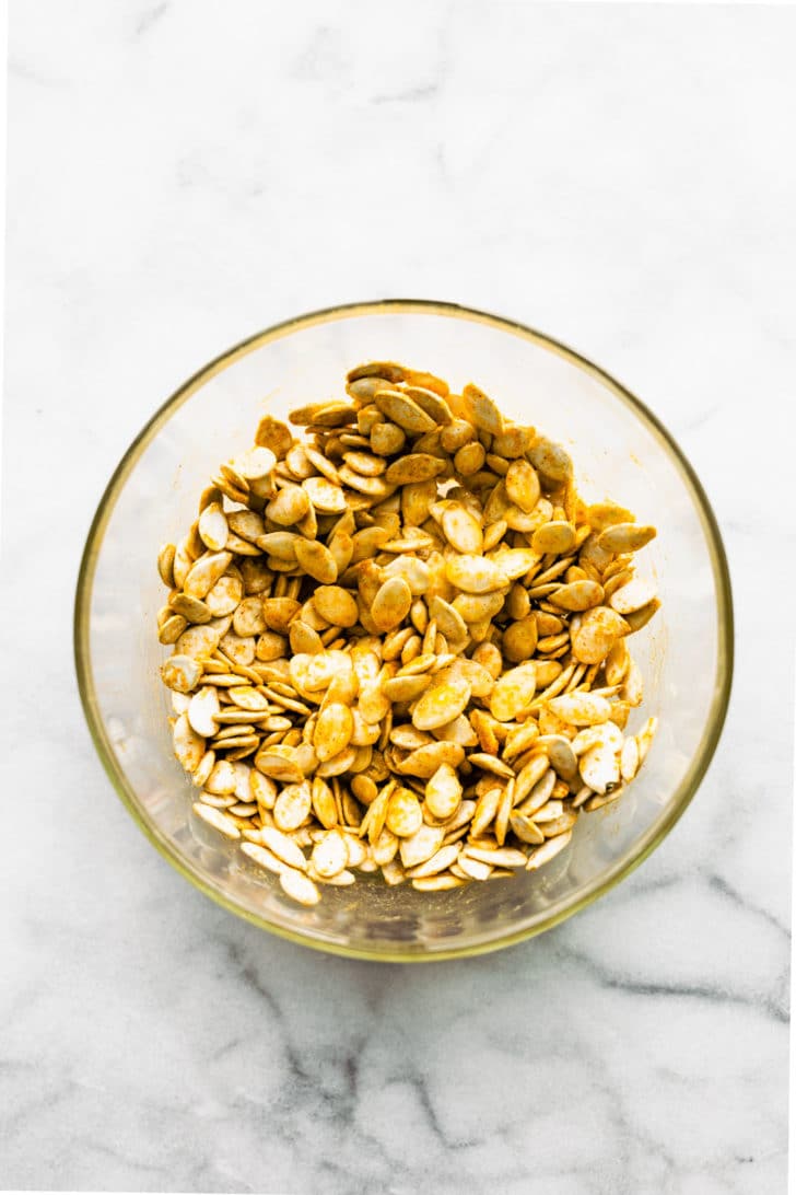 A clear glass bowl filled with raw pumpkin seeds tossed in butter and seasonings.