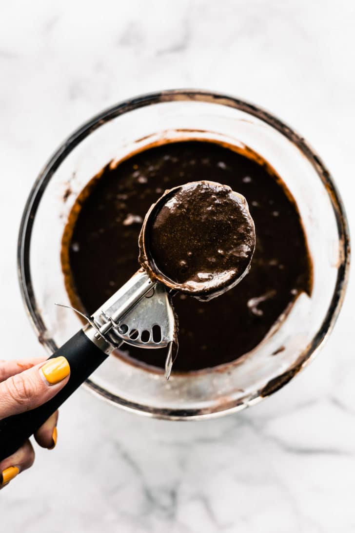 A scoop holding a full scoop of gluten free chocolate cupcake batter over a bowl of batter