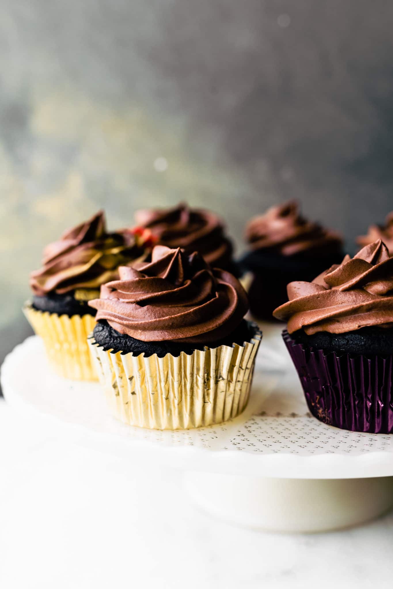 Vegan chocolate cupcakes in gold and purple liners on a white cake plate.