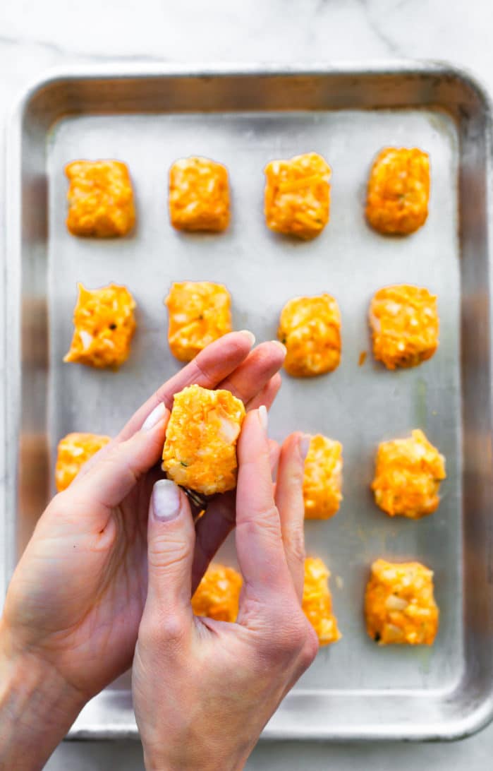 two hands holidng a sweet potato tot above a tray full of tots