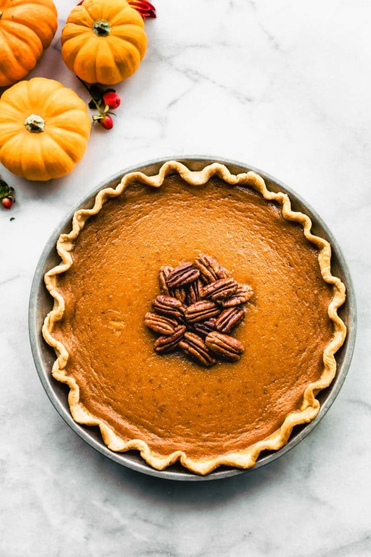 Overhead view of gluten free pumpkin pie topped with pecans in crimped edge pie crust in a metal pie plate, mini pumpkins to upper left corner of image.