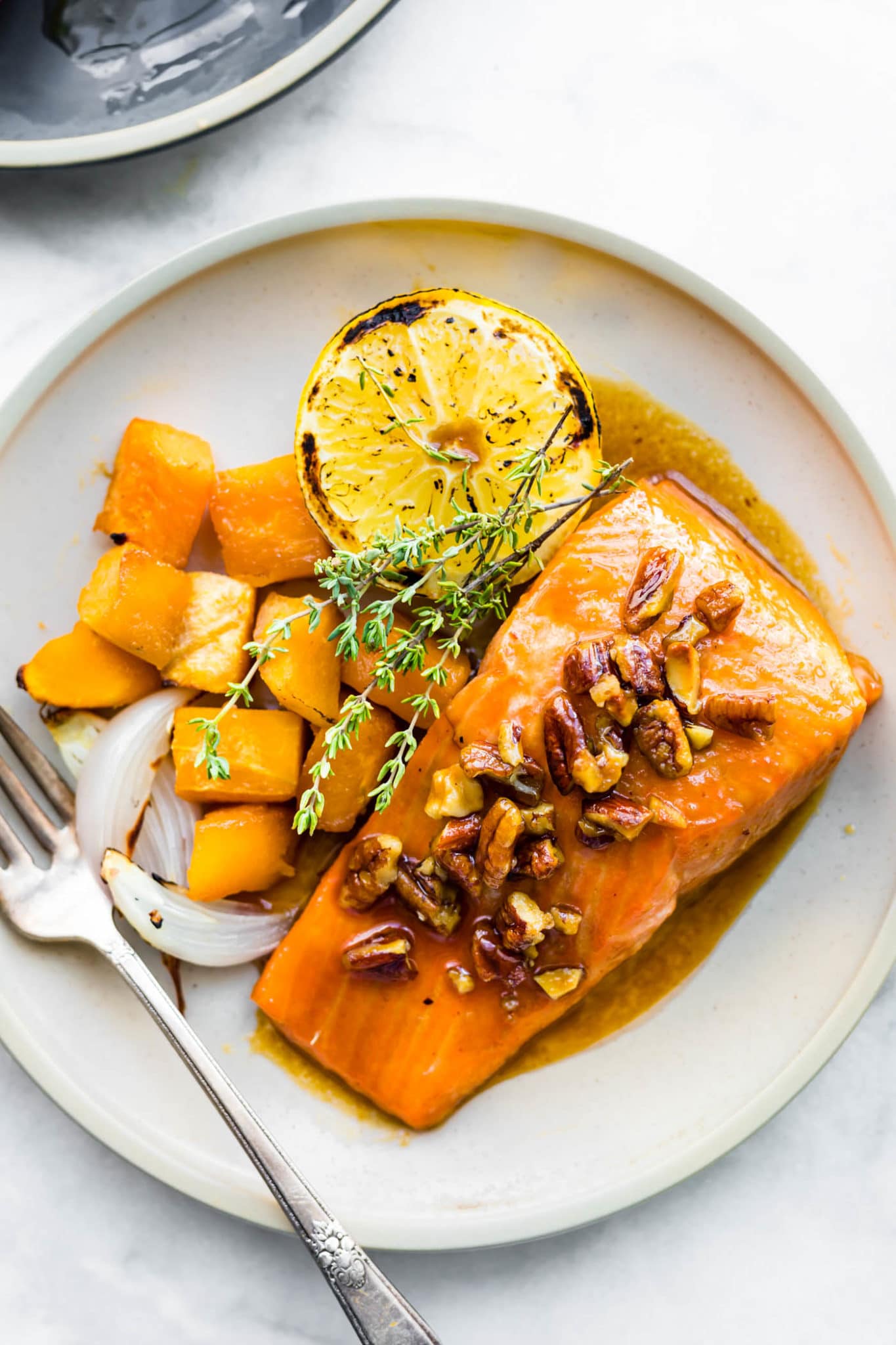 close up image of maple bourbon glazed salmon topped with candied pecans on a cream speckled plate with side of roasted butternut squash and slightly charred edge lemon half.