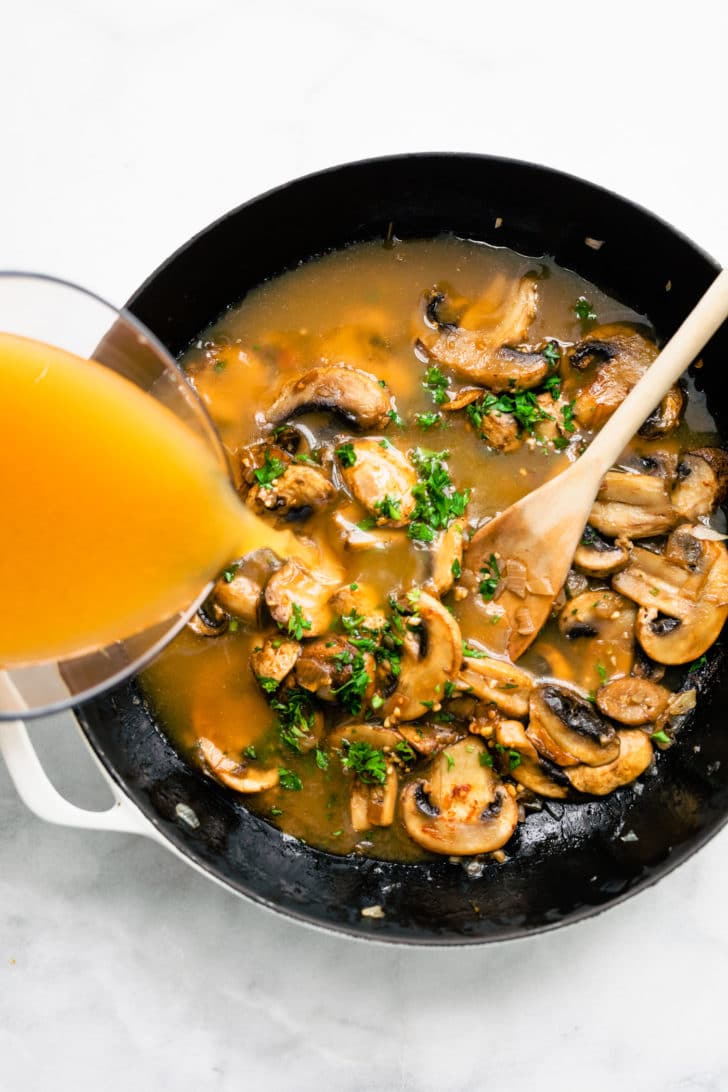 broth being poured into a pan with mushrooms and herbs for vegan mushroom gravy