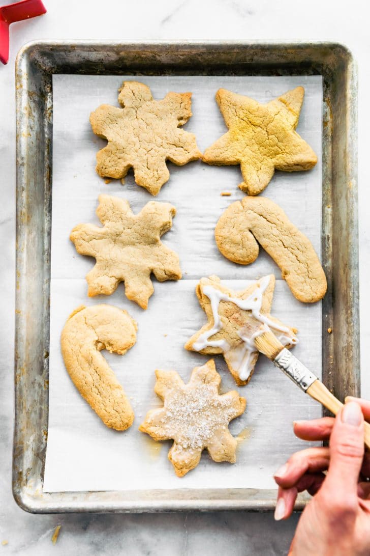 Overhead view of gluten free sugar cookie cut out on a parchment lined baking sheet, a hand holding a paint brush brushing on icing.