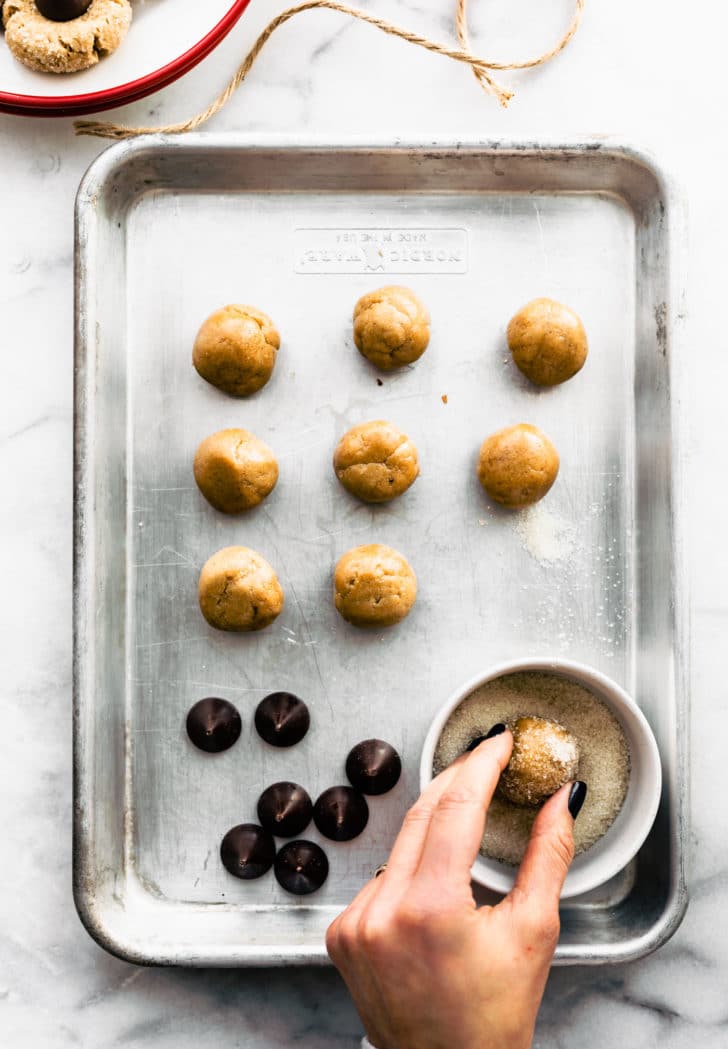 ingredients for peanut butter blossom cookies on a baking sheet with cookie dough balls arranged in a line, and a hand rolling a cookie dough ball in sparkling cane sugar