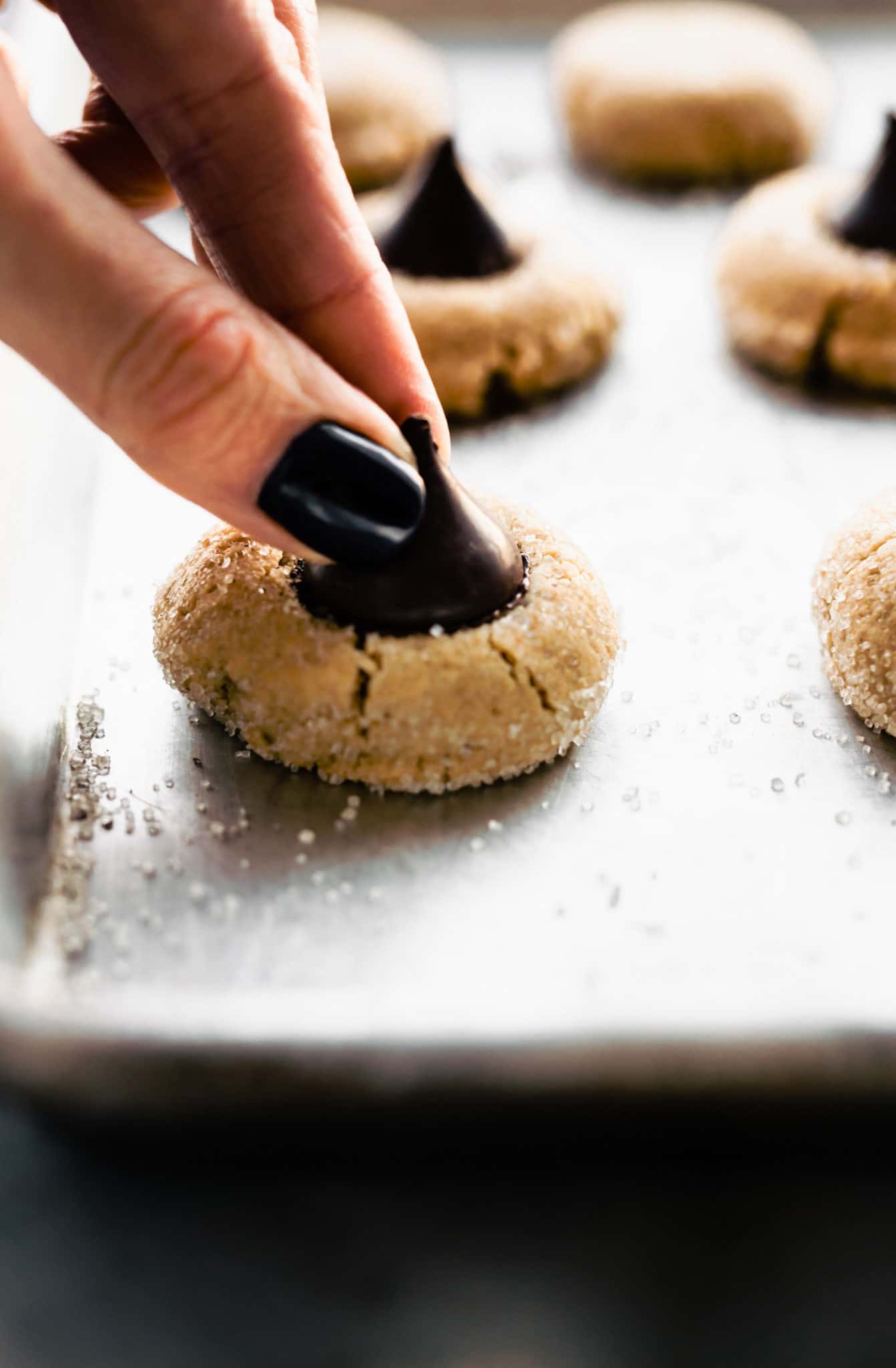 two fingers pushing a dark chocolate kiss into a peanut butter blossom cookie on a baking sheet