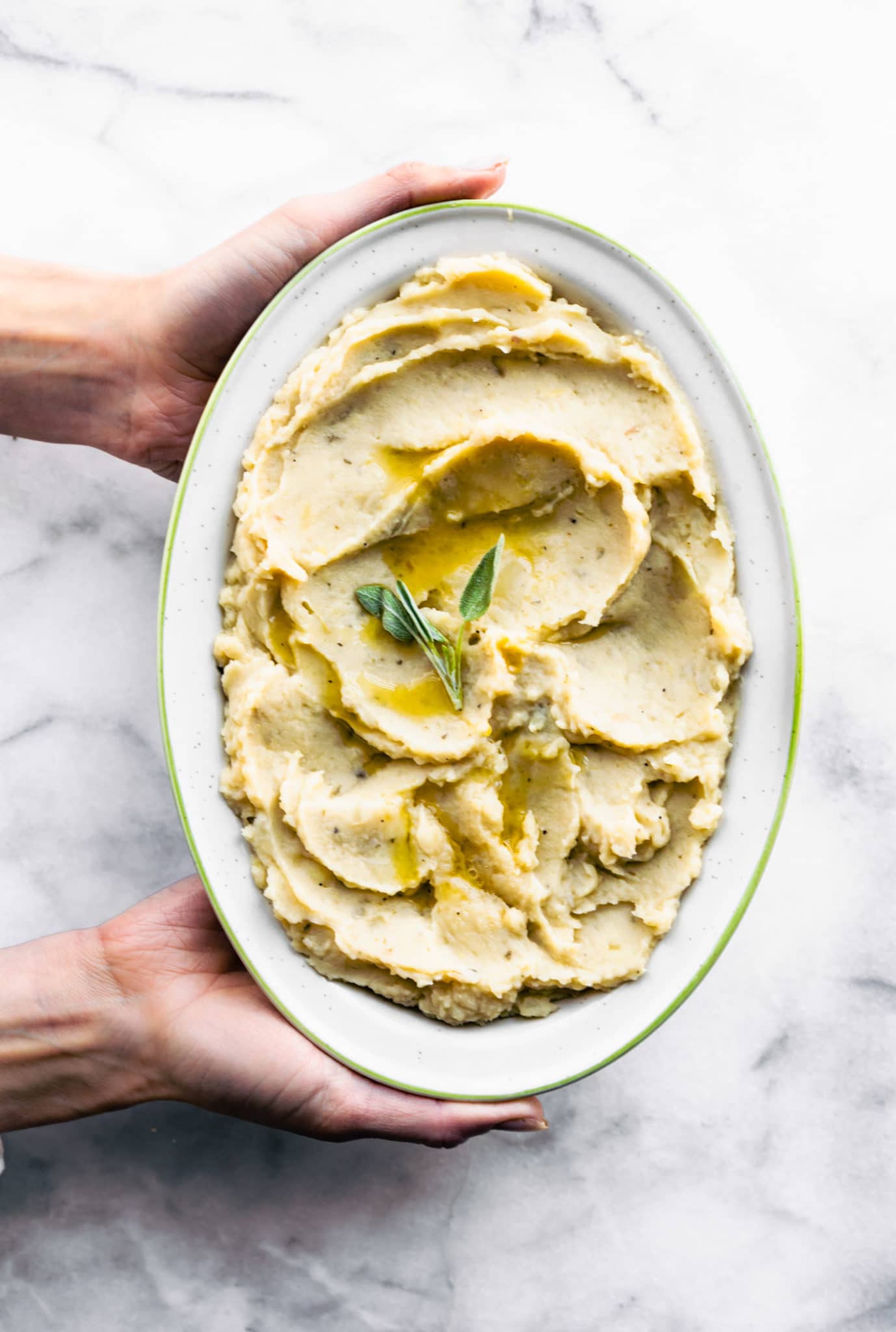 Two hands holding a platter of slow cooker dairy free mashed potatoes topped with fresh herbs